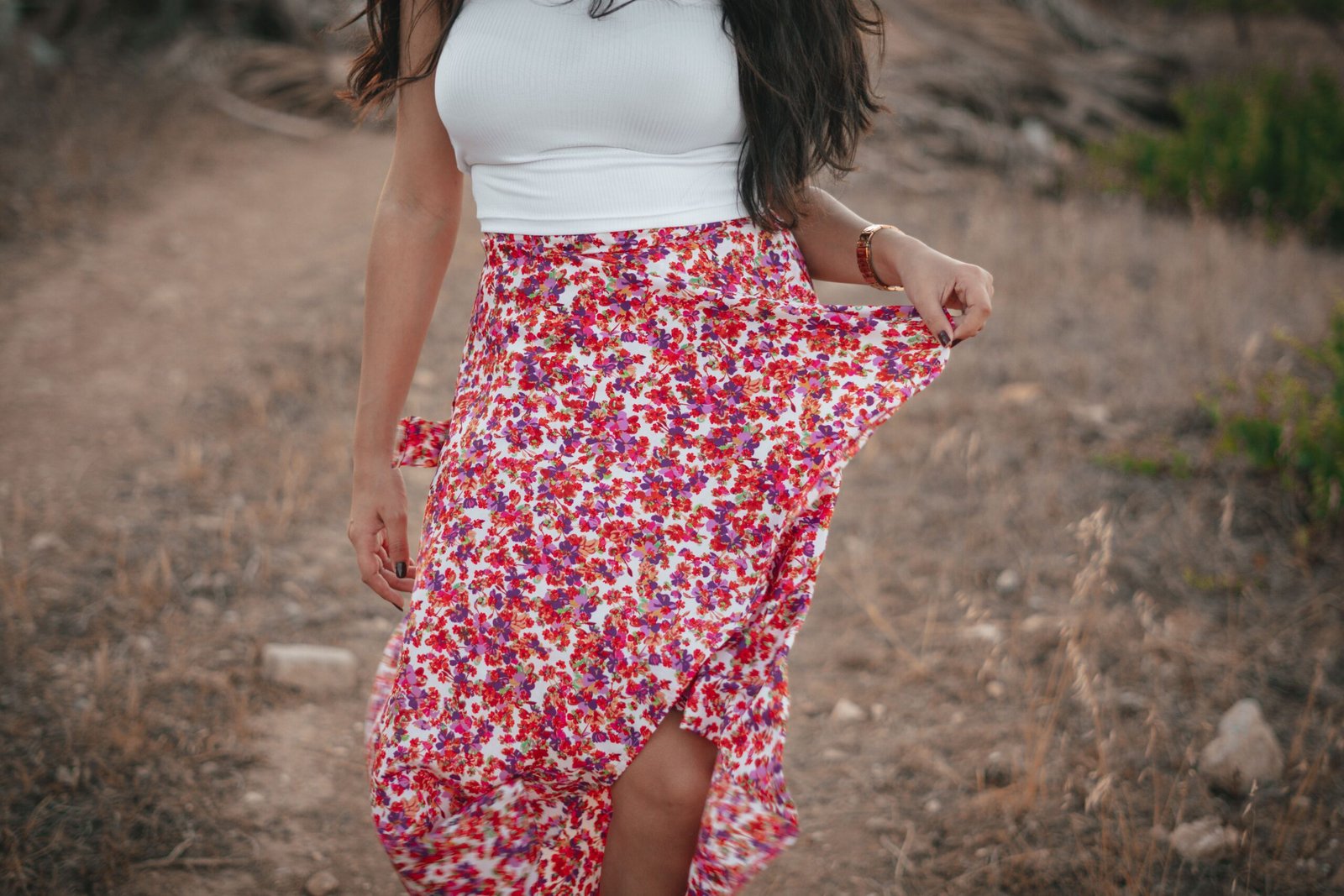 An adult woman in a floral skirt and white top walking in a dry outdoor setting.